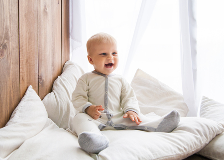Toddler smiling cheerfully and looking away sitting on white feather with pillows near wooden wallの写真素材