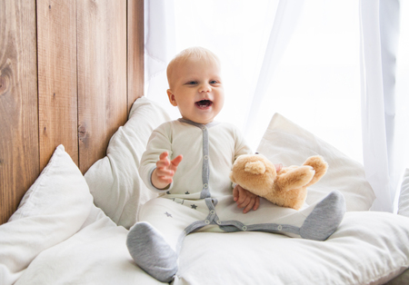 Smiling toddler sitting and holding teddy bear near wooden wall and window with white curtain.の写真素材