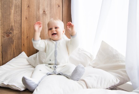 Toddler smiling cheerfully and looking away sitting on white feather with pillows near wooden wallの写真素材