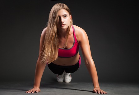 Young girl in sport outfit making push-up on hands and looking at camera on plain black background.の写真素材
