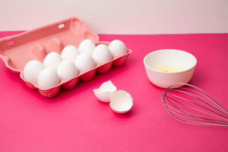 Box of eggs near bowl of yolk and whisk on pink table