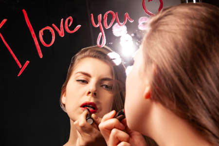 Lovely female looking in mirror with inscription I Love You and applying red lipstick while doing makeupの写真素材
