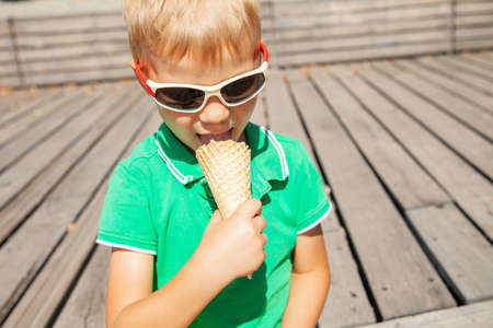 Adorable blond haired little boy in fashionable sunglasses biting yummy vanilla ice cream cone while spending summer day in parkの写真素材
