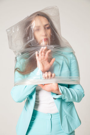 Full body trendy young female in blue suit covered with transparent plastic bag standing against...