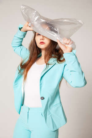 Young female model in stylish blue suit looking at camera and removing plastic bag from head against gray backgroundの写真素材
