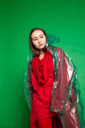 Calm female wearing transparent plastic raincoat and red clothes standing on green background in studioの写真素材