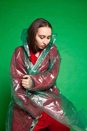 Calm female wearing transparent plastic raincoat and red clothes standing on green background in studioの写真素材