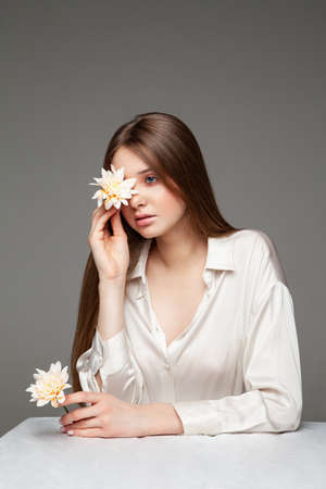 Young female with long brown hair covering eye with natural flower and looking away while sitting at table against gray backgroundの写真素材