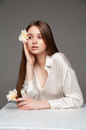 Young female with long brown hair covering eye with natural flower and looking away while sitting at table against gray backgroundの写真素材