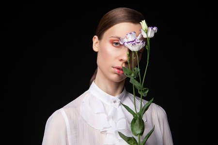 Pretty young woman in elegant white blouse touching face with natural flowers and looking away against black backgroundの写真素材