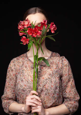 Side view of young female in fred blouse hiding face behind natural flowers against black backgroundの写真素材