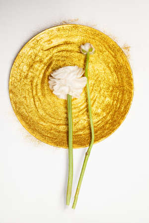 Top view of natural fresh peonies placed on round plate covered with glittering golden paint on white backgroundの写真素材
