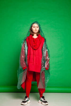 Calm female wearing transparent plastic raincoat and red clothes standing on green background in studioの写真素材