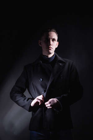 Serious confident young male in black coat looking away while standing under light in dark studio with black wall and shadowの写真素材