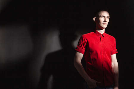 Serious confident young male in bright red shirt looking away while standing under light in dark studio with black wall and shadowの写真素材