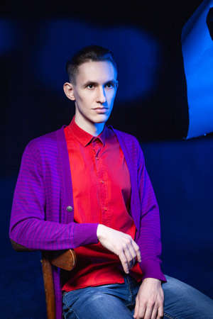 Young male in bright red shirt and purple blouse looking away while sitting on chair in dark studio with black and blue wall and shadowの写真素材