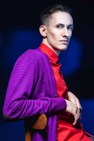 Young male in bright red shirt and purple blouse looking away while sitting on chair in dark studio with black and blue wall and shadowの写真素材