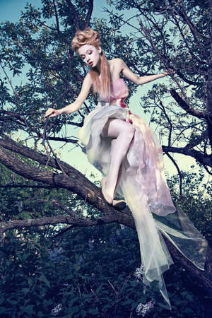 Lovely young lady wearing elegant white dress with roses standing on the tree in woodの写真素材