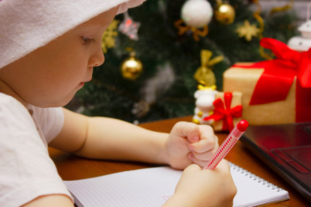 A child boy in a hat of santa claus writes a letter to santa claus on the background of an elegant christmas tree and a computerの写真素材