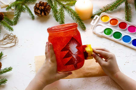 the boys hands make a candlestick from a glass jar painted with red paint with a silhouette of a Christmas tree, step by step instructions, step 8.の写真素材
