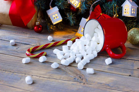 cozy winter card with scattered marshmallows from red mug on background of christmas decor, christmas decor with garlands and gift on wooden backgroundの写真素材