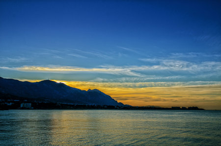 sunset on the sea bright sky with clouds on the left you can see the mountains no longer lit by the sun and the orientation is landscape there is a place for textの写真素材