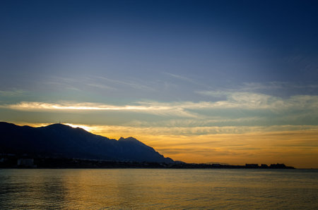 sunset on the sea bright sky with clouds on the left you can see the mountains no longer lit by the sun and the orientation is landscape there is a place for textの写真素材