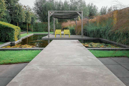 Two yellow chair chairs under the pergola which is surrounded by a pool of water to enjoy the sun in a corner of a beautiful garden.の写真素材