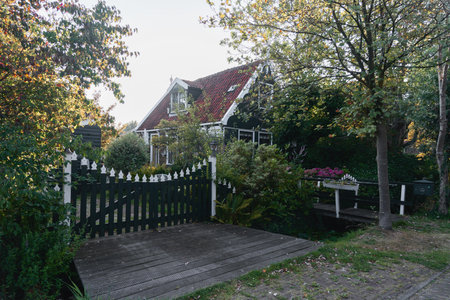 A rustic countryside house featuring a small wooden bridge over a pond, bathed in warm sunlight and surrounded by trees and greenery.の写真素材