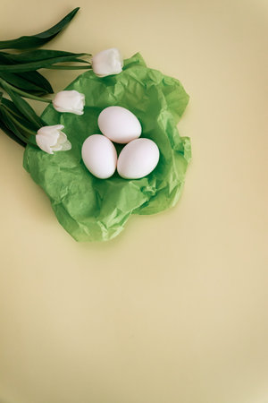 white tulips with colorful eggs in a nest of green silk on a pale yellow background