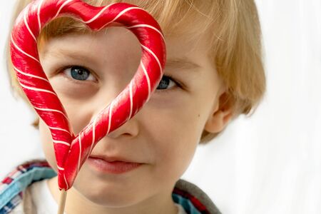 Cute little boy in with candy red lollipops in heart shape, white background. Beautiful kid eat sweets. Valentines day, love concept.の写真素材
