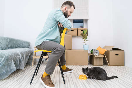 Man sits on yellow chair in front of his cat with the background of moving boxes, while moving into new apartment.の写真素材
