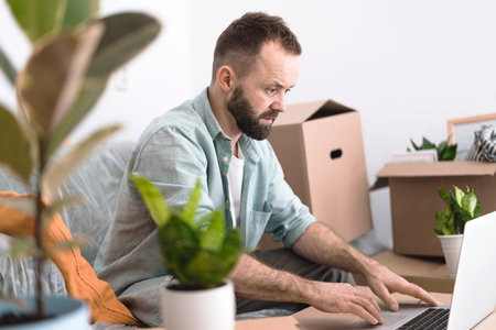 A mature man sitting in a messy room with cardboard boxes and plants, using laptop. A moving...