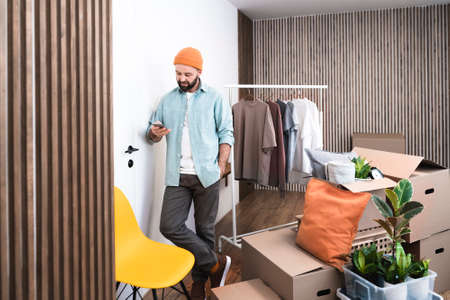 Mature man typing text message on his smartphone, unpacking things from boxes while moving in new unfurnished apartment.の写真素材