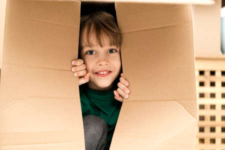 Boy hiding in inside a huge cardboard box. He is playing and peeking through a hole in box. Kid is happy about moving into a new home.の写真素材