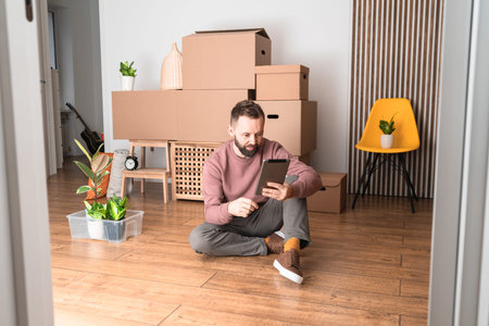 Mature man using his tablet, unpacking things from boxes while moving in new unfurnished apartment.