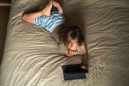 Boy lying on the bed using digital tablet computer playing games or watching cartoons at home. Child internet safety concept. Kid using electronic equipment. Parental control. Online education.の写真素材