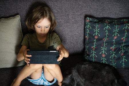 Boy sitting on sofa using digital tablet computer playing games or watching cartoons at home. Child internet safety concept. Kid using electronic equipment. Parental control. Online education.の写真素材