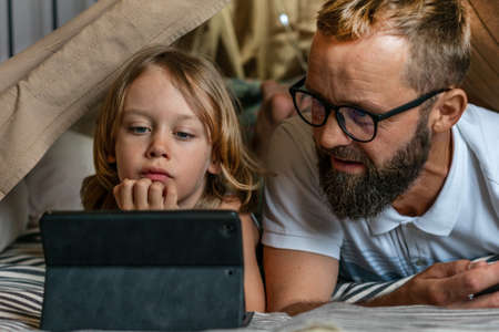 Portrait of a 6 year old boy and his father having fun playing in teepee tent. Father and son using digital tablet watching cartoons or playing computer games lying in kid tent at home.の写真素材