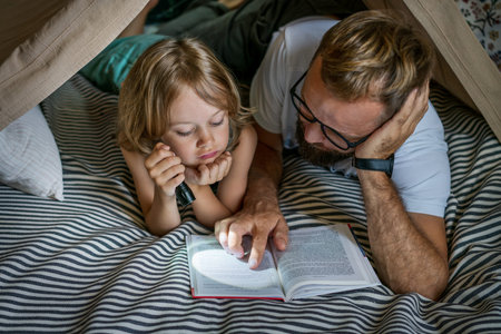 Portrait of a 6 year old boy and his father reading a book in teepee tent. Father and son with...