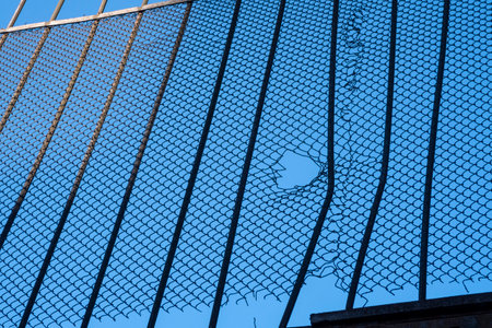 Damaged metal grid fence with hole against clear blue sky.の写真素材