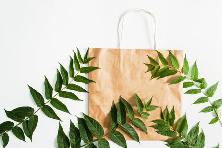 Paper bag and green leaves isolated on white background. Ready-made eco shopping concept. Top view....