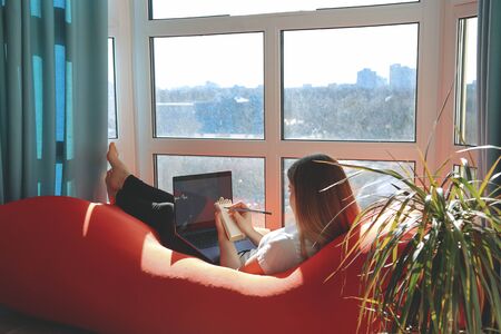Work at home. Beautiful young smiling woman working on laptop while sitting in a big comfortable chair at home next to the windowの写真素材