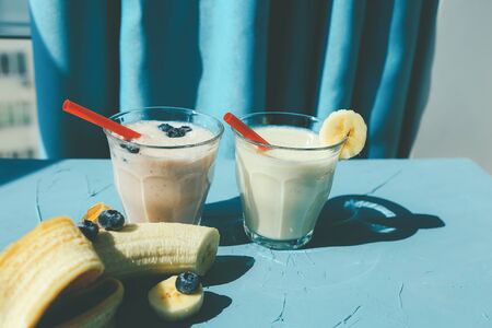 Two Glasses of banana blueberry milkshake or smoothie with drinking straw and banana on blue backgroundの写真素材