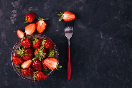 One strawberry on a red fork and in the crystal bowl of the black rustic background. Top view. Vertical. Diet minimal concept.の写真素材