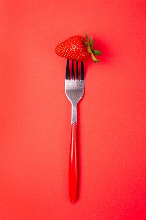 One strawberry on a red fork in the center of the red background. Top view. Vertical. Diet minimal concept.の写真素材