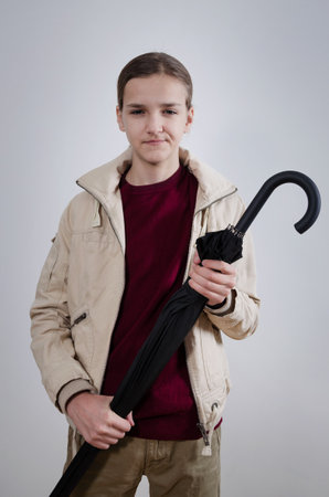 Stylish handsome teenager model holding a folded black umbrella while standing against isolated grey background.の写真素材