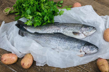 Raw peeled trout with potatoes and parsley on the rustic wooden table. Fresh fish wapped in parchment paper and vegetables and parsley on wooden background. Tasty fishes preparing for lunch.の写真素材