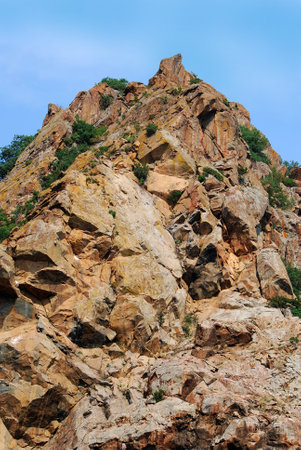 Scenic view of single mountain with the bushes and grass. A picturesque view of a lonely rock with bushes and grass against the backdrop of a clear sky. Bottom view of a rock wall in good weather.の写真素材