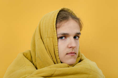 A portrait of the teenager boy covered with blanket looking at camera against white background. The young man wrapped himself in a warm blanket basks on a white background.の写真素材
